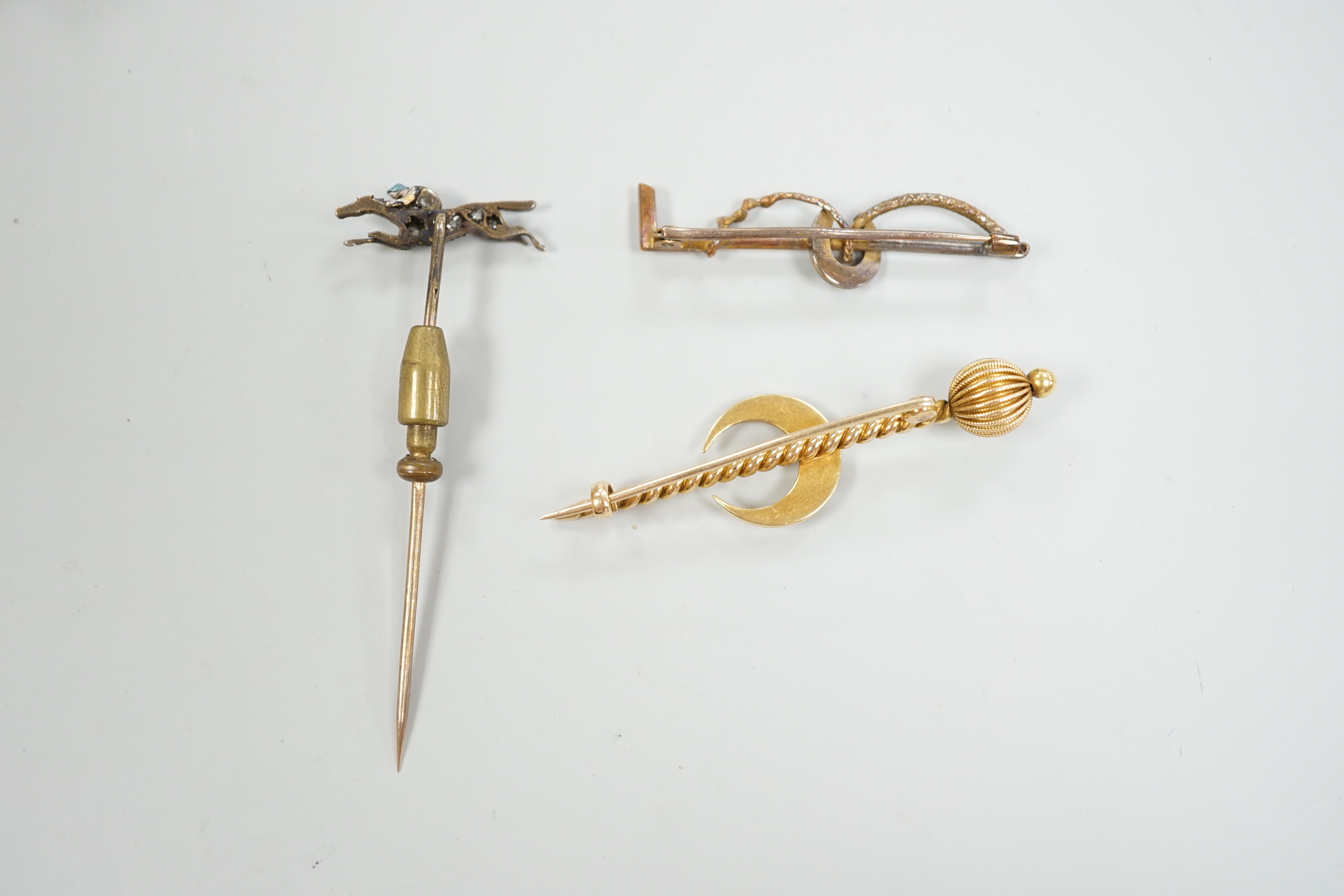 An Edwardian enamel and diamond set 'horse and jockey' stick pin, 63mm, a yellow metal and seed pearl set 'crescent and staff, brooch and a 'riding crop and horseshoe' brooch.